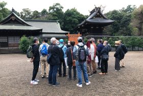 神社本殿（右側）拝殿（左側）の前で観光ボランティアガイドの説明を聞く